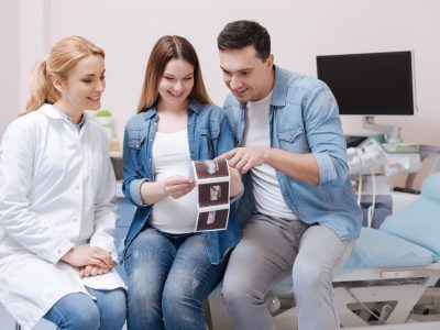 Happy doctor demonstrating ultrasonic scan to the patient at work Sharing delightful vibes with patient. Optimistic smiling professional doctor working in the hospital while having conversation with young couple and showing digital fetus scan